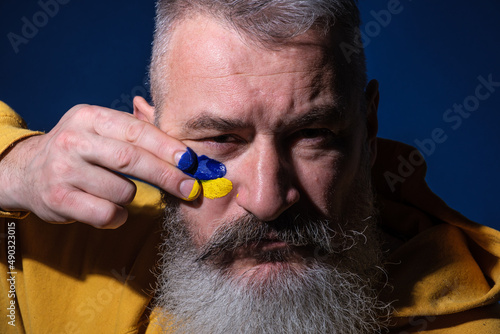 Mature bearded man dressed yellow hoodie draws Ukrainian flag on his face, protest against war