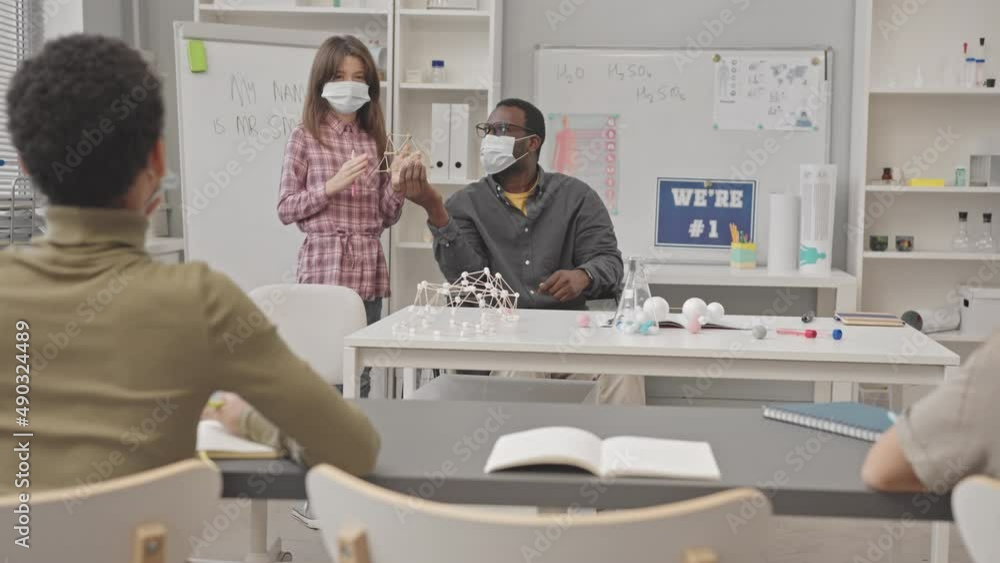 Slowmo of elementary school female student in face mask making report on molecular compounds standing next to African American Chemistry teacher holding plastic molecular model Slowmo of elementary school female student in face mask making report on molecular compounds standing next to African American Chemistry teacher holding plastic molecular model