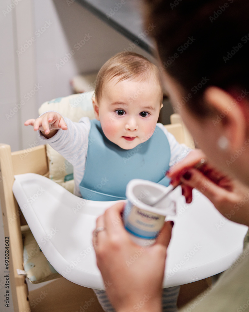 Mother giving a yogurt to her son sitting in the highchair Mother giving a yogurt to her son sitting in the highchair