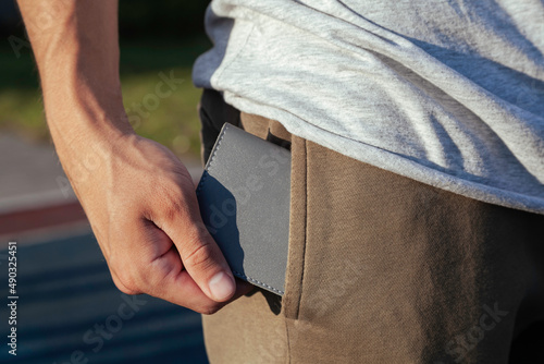 A young man in close-up sportswear puts a small and comfortable gray leather wallet in his back pocket against the background of the sky and trees. Training on the playground. 