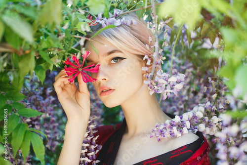 A beautiful girl with blonde hair in a traditional Chinese black and red dress. Portrait in a blooming wisteria garden, around flowers. The girl holds a branch of red flower in her hand. Close up face