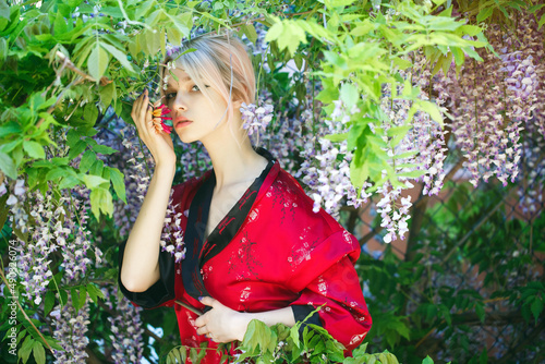 A beautiful girl with blonde hair in a traditional Chinese black and red dress. Portrait of an attractive woman in a blooming wisteria garden. The girl holds a branch of red flower in her hand.