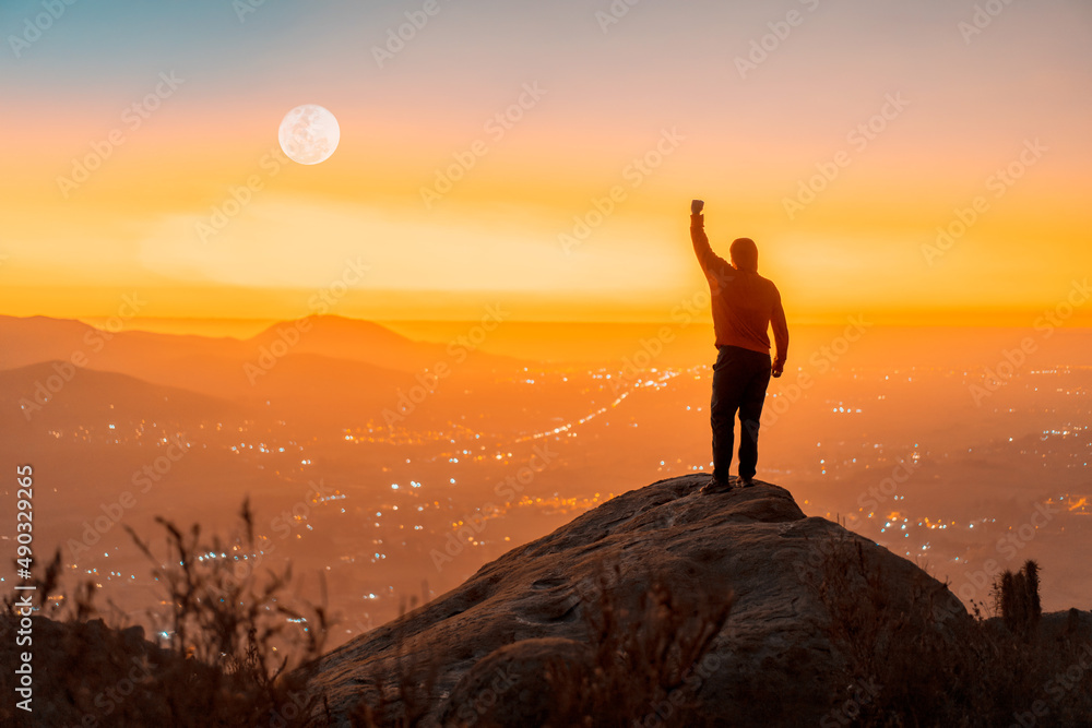 person standing on the top of the mountain with hand up, back view, over the city at sunset person standing on the top of the mountain with hand up, back view, over the city at sunset