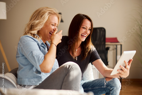 Two young women sitting on sofa use a digital tablet and laughing