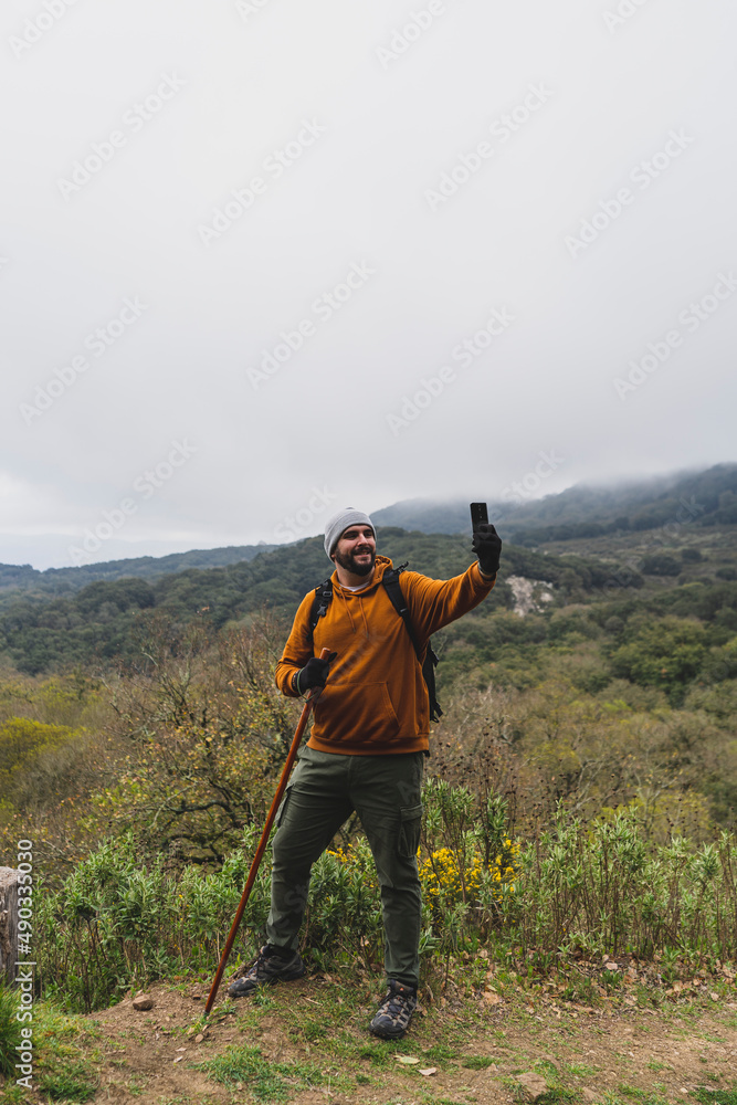 Chico con barba muy alto delante de la sierra malagueña realizando fotos a si mismo tipo selfie con el movil