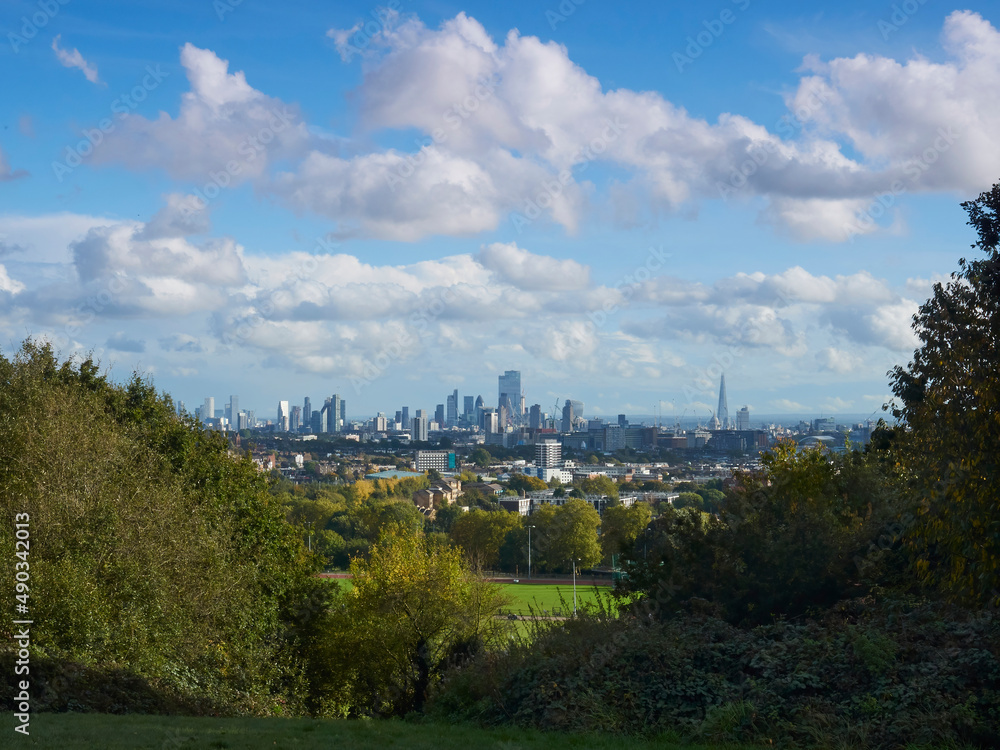 London from Parliament Hill Fields, trees and bushes ahead of the famous vista of skyscrapers and storied buildings, under a bright autumnal sky.