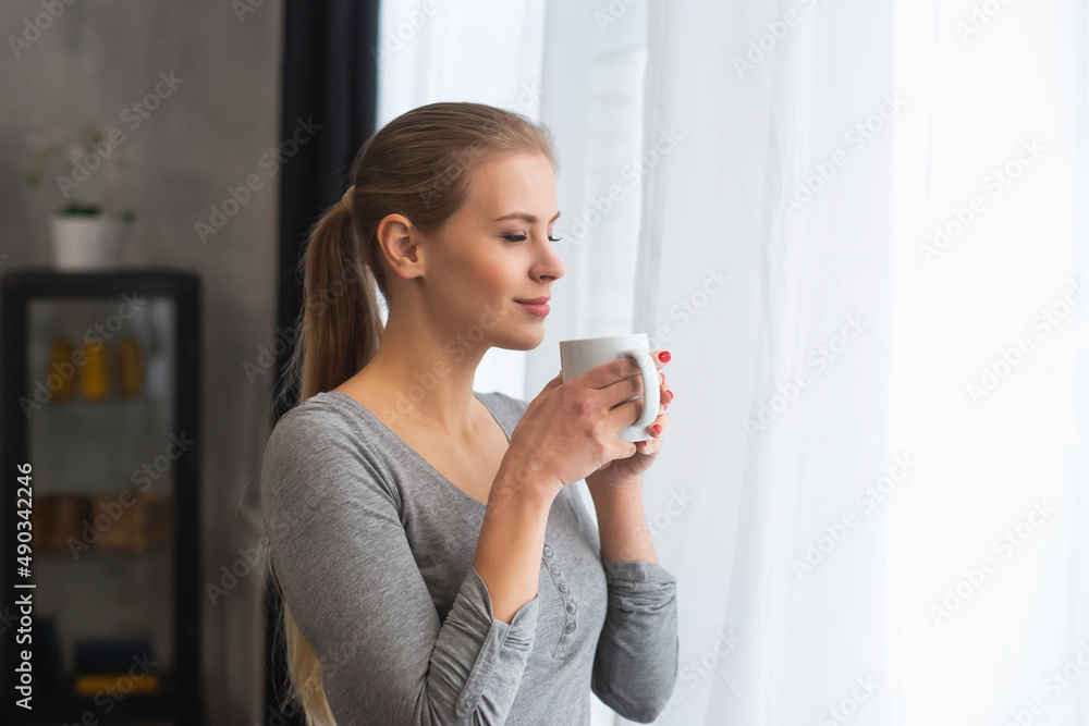 Young teenage girl is standing in front of the window and looking into it. Blond woman at home. Young teenage girl is standing in front of the window and looking into it. Blond woman at home.