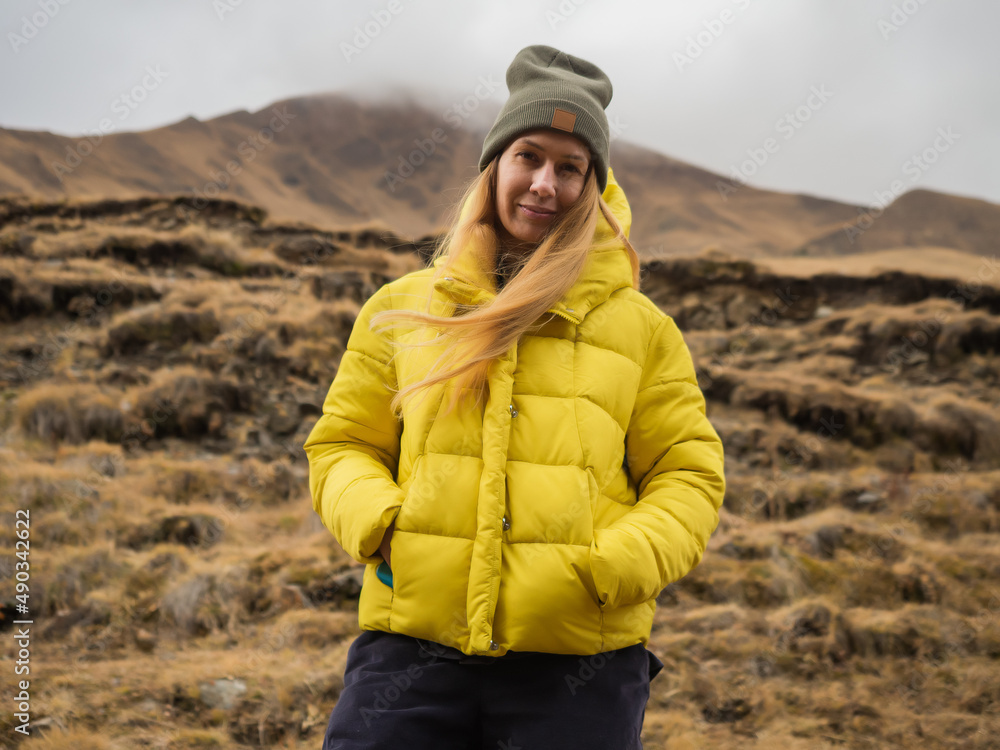 A woman tourist in a quilted yellow jacket stands smiling against the backdrop of nature in the mountains A woman tourist in a quilted yellow jacket stands smiling against the backdrop of nature in the mountains