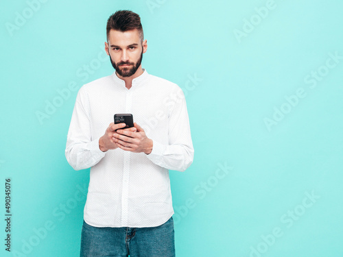 Handsome confident model.Sexy stylish man dressed in shirt and jeans. Fashion hipster male posing near blue wall in studio. Holding smartphone. Looking at cellphone screen. Using apps © halayalex