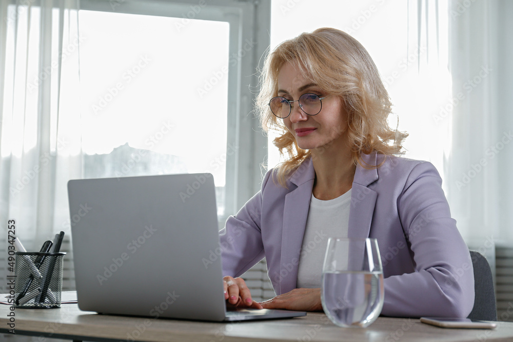 Portrait of adult blonde woman wearing lilac jacket and glasses sitting at her workplace. Smart looking businesswoman working on a laptop in the office. Background, copy space, close up. Portrait of adult blonde woman wearing lilac jacket and glasses sitting at her workplace. Smart looking businesswoman working on a laptop in the office. Background, copy space, close up.