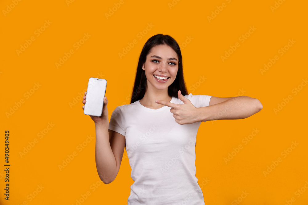 Young beautiful brunette woman with long hair wearing white t-shirt, posing with a phone over isolated yellow background. Portrait of female model showing blank screen cellphone. Close up, copy space. Young beautiful brunette woman with long hair wearing white t-shirt, posing with a phone over isolated yellow background. Portrait of female model showing blank screen cellphone. Close up, copy space.