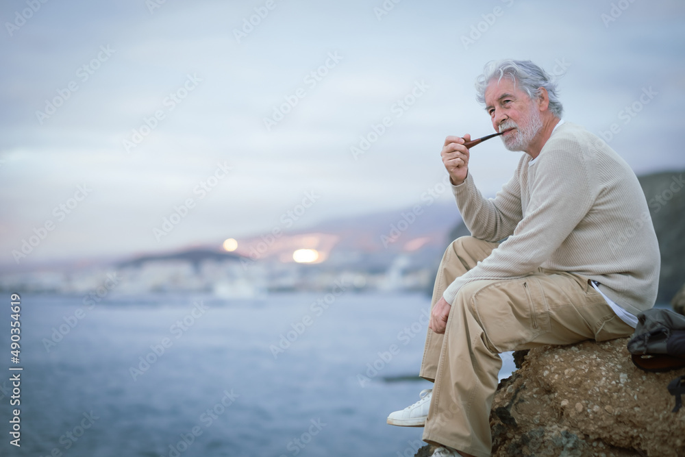 Adult old senior bearded man resting during excursion at sea smoking pipe, looking at horizon over water. Caucasian elderly white-haired male enjoying free time, vacation and nature in seascape.