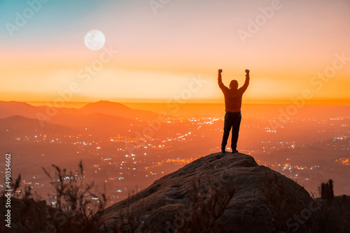 person silhouette on the top of the mountain with hands raised, back view, sunset background
