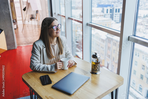 A modern woman of Slavic appearance, a manager in a light jacket and glasses, a girl with a smile on her face, sits at a table in a cafe and drinks tea. Remote work © Дмитрий Ткачук