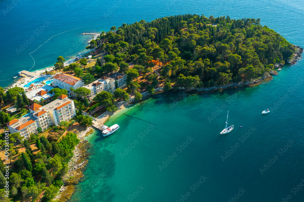 Buildings with red rooftops on peninsula with green forest near Adriatic sea. Yachts drifting on calm water surface near Croatian town. Aerial panorama