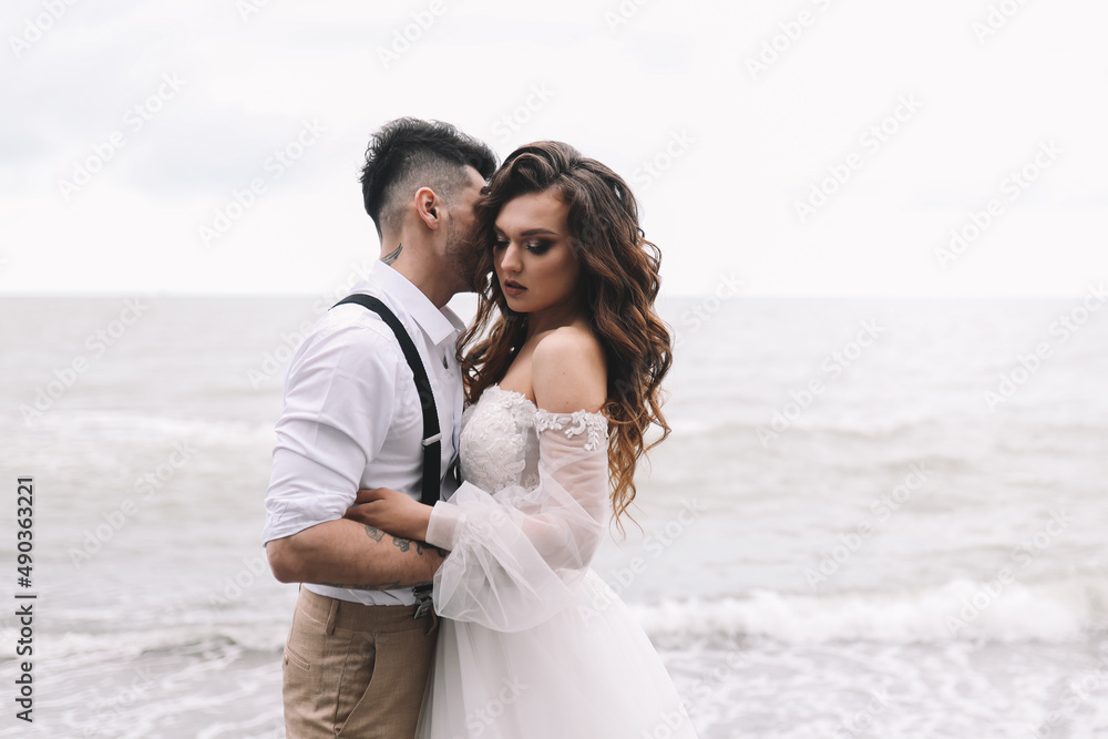 The bride and groom in wedding dresses hug and walk together along the sea in nature on an outdoor trip. Husband and wife in love spend their honeymoon. Selective focus The bride and groom in wedding dresses hug and walk together along the sea in nature on an outdoor trip. Husband and wife in love spend their honeymoon. Selective focus