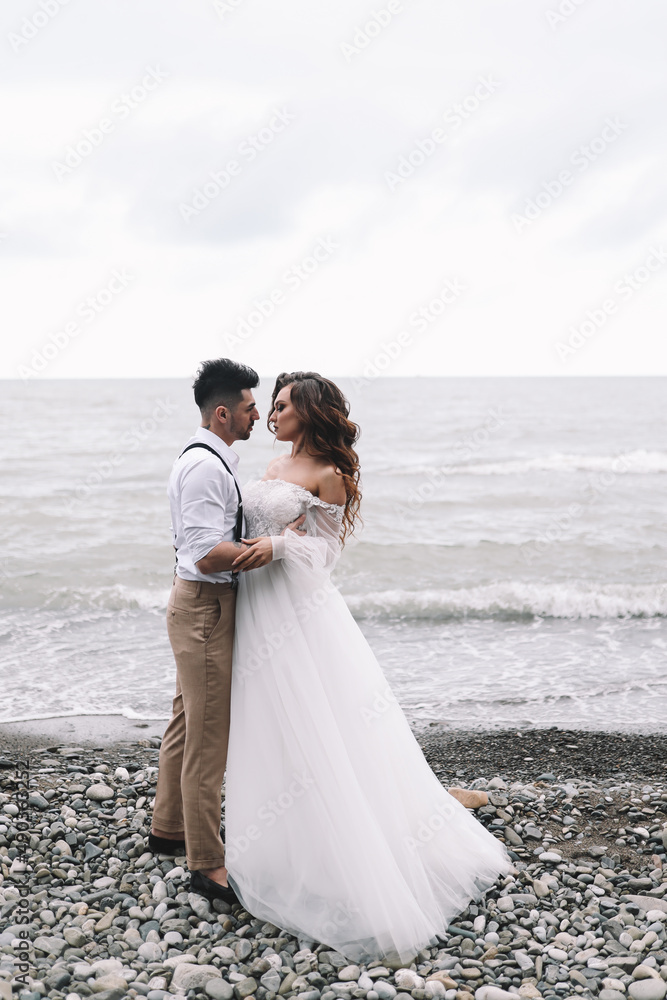 The bride and groom in wedding dresses hug and walk together along the sea in nature on an outdoor trip. Husband and wife in love spend their honeymoon. Selective focus The bride and groom in wedding dresses hug and walk together along the sea in nature on an outdoor trip. Husband and wife in love spend their honeymoon. Selective focus