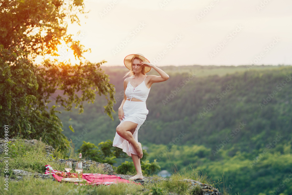 Attractive young woman in white dress and straw hat in the mountain on a summer picnic. Summer vacation and picnic. Attractive young woman in white dress and straw hat in the mountain on a summer picnic. Summer vacation and picnic.