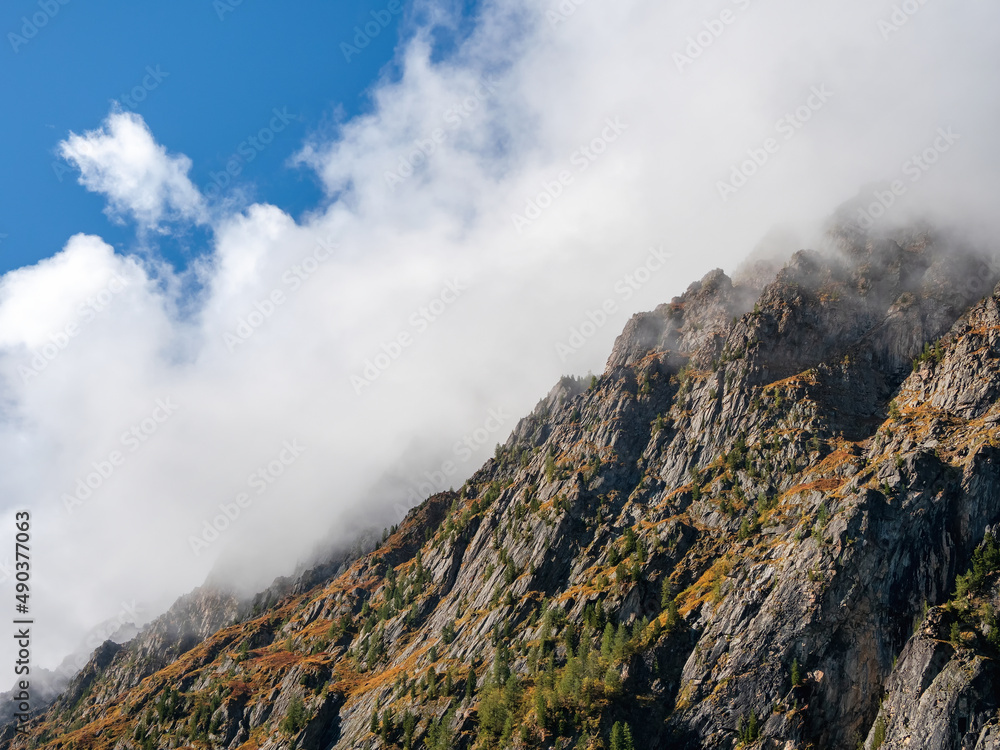 Crest of the mountain under white low clouds. Ghostly atmospheric view to big cliff in cloudy sky. Low clouds among giant rocky mountains.Natural background with high mountains, a steep mountain slope