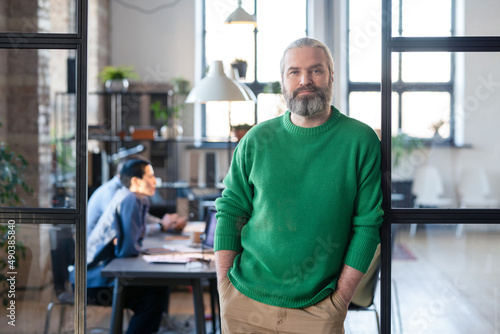 Portrait of mature bearded manager in casual clothing leaning on the glass wall and looking at camera while having meeting with colleagues