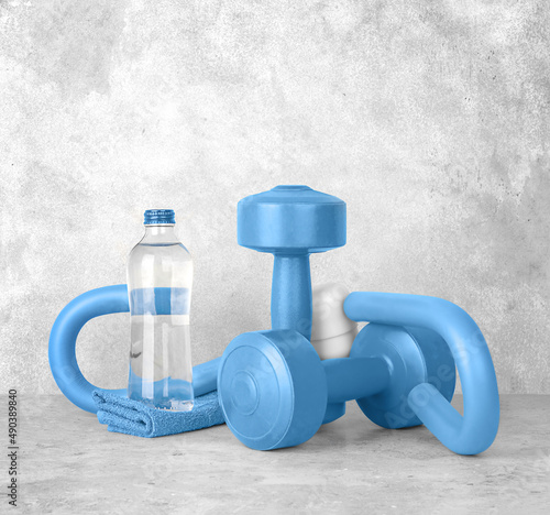 Set of fitness equipment on a gray concrete background, front view, close-up. Blue dumbbells, s-shaped leg exercise machine, glass bottle of water, towel. Home workout. Fitness and activity.
