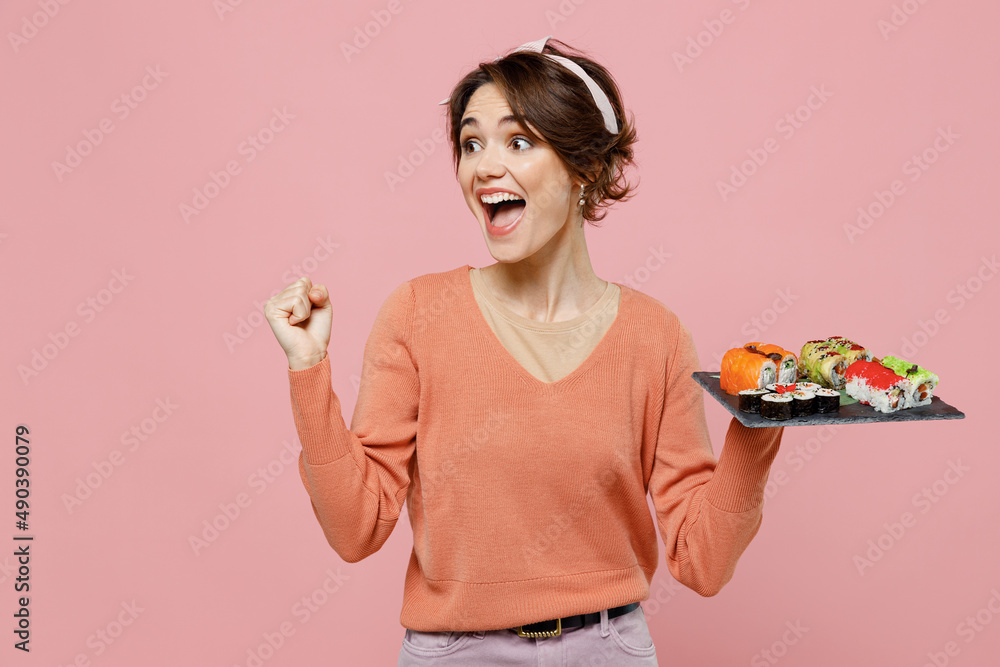 Young woman in sweater hold makizushi sushi roll served on black plate traditional japanese food do winner gesture celebrate clench fists say yes look aside isolated on plain pastel pink background Young woman in sweater hold makizushi sushi roll served on black plate traditional japanese food do winner gesture celebrate clench fists say yes look aside isolated on plain pastel pink background