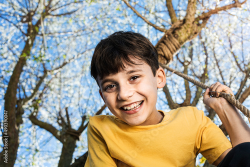 Beautiful happy and carefree boy smiling while climbing on a tree swing joyful for the arrival of spring. Tree blossoming in spring and a blue sky in the background. spring solstice photo