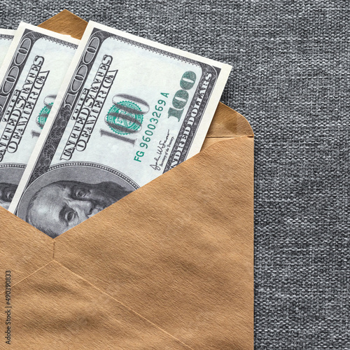 A square photo of hundred-dollar bills in an envelope on a gray fabric background. The concept of a bribe of American banknotes in an envelope.