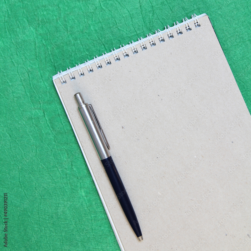 White and gray notepad sheet with spiral with pen against the background of green fabric. Concept of analysis, study, attentive work. Stock photo with empty place for your text and design. Square White and gray notepad sheet with spiral with pen against the background of green fabric. Concept of analysis, study, attentive work. Stock photo with empty place for your text and design. Square