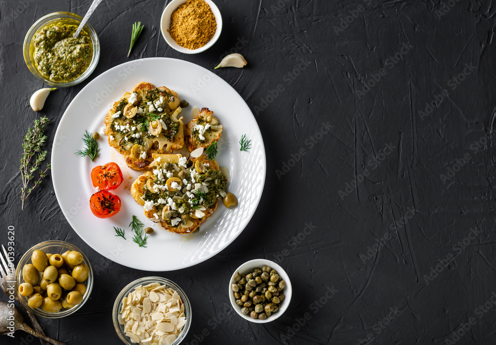 Cauliflower steak with spices, chimichurri sauce, almond flakes, olives, fried cherry tomatoes and capers on a white plate. Dark background. Copyspace.