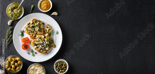 Cauliflower steak with spices, chimichurri sauce, almond flakes, olives, fried cherry tomatoes and capers on a white plate. Dark background. Copyspace. Banner.