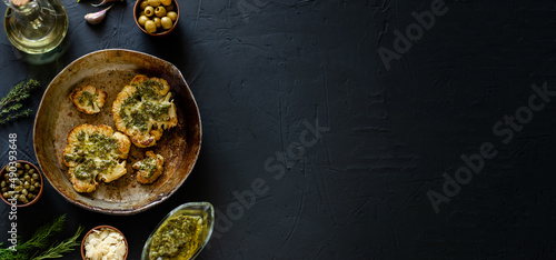 Cauliflower steak with spices lies in a frying pan. Olive oil, chimichurri sauce, herbs, various spices side by side. Dark background. Vegetarian food. Banner, copyspace.