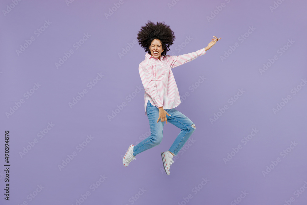 Full body singer musician expressive young woman of African American ethnicity 20s in pink striped shirt jump high play guitar gesture isolated on plain pastel light purple background studio portrait.