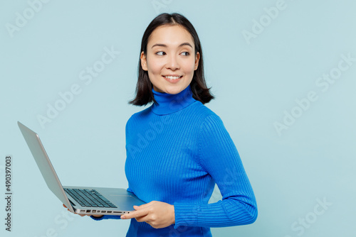 фотографія Calm fancy fascinating young woman of Asian ethnicity 20s years old wears blue shirt hold use work on laptop pc computer looking aside isolated on plain pastel light blue background studio portrait