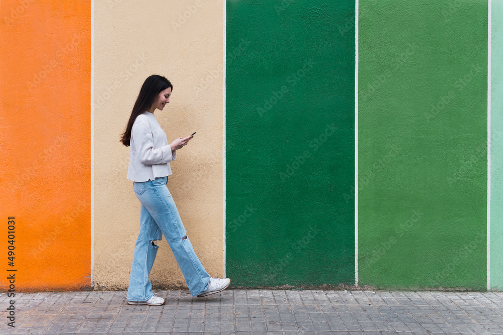 Asian woman walking while using her smartphone against colorful wall