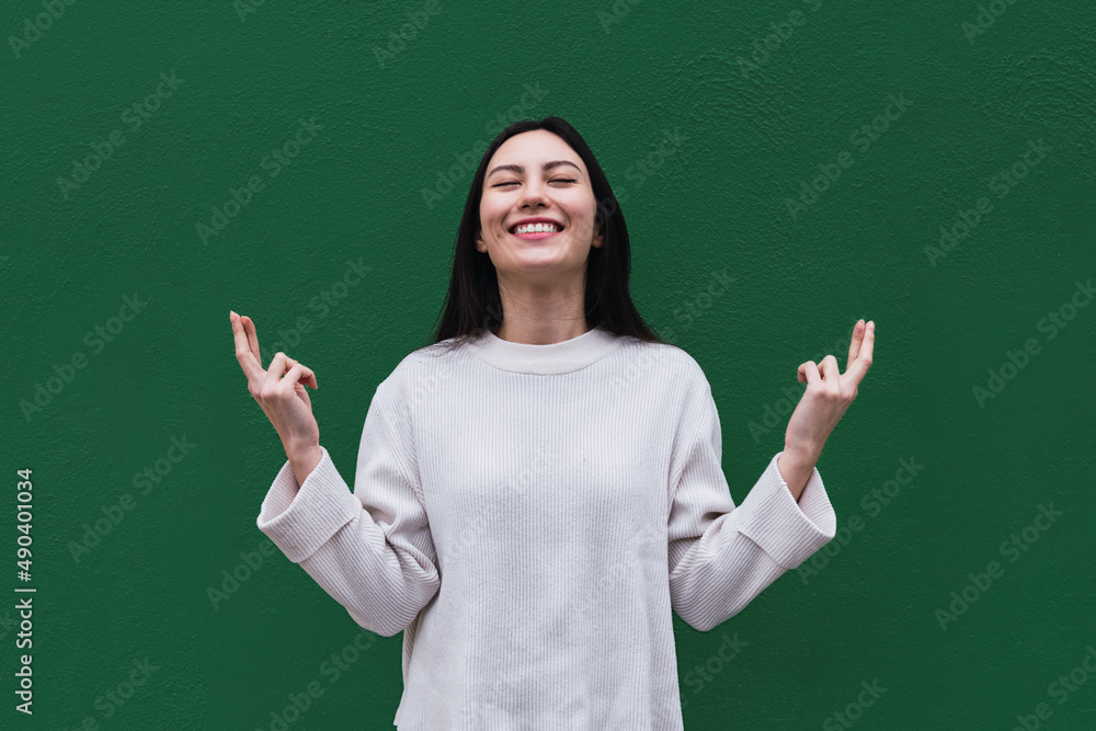 Asian woman smiles broadly with eyes closed, keeps fingers crossed, hope for good luck during exam, isolated over green background with copy space for your text