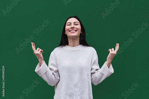 Платно Asian woman smiles broadly with eyes closed, keeps fingers crossed, hope for goo