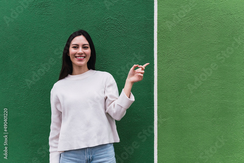 Asian woman feeling happy and smiling casually, looking to an object or concept held on the hand on the side isolated against green wall © Frenchiebuddha
