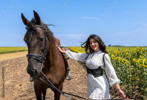Confident brunette standing in agriculture field with brown horse. She is wearing traditional white dress and black leather corset. Sunny summer day. Rural scene.