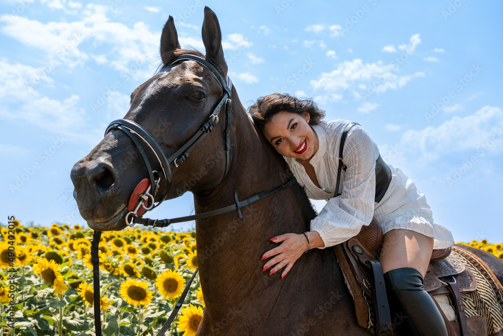 Obraz premium Cheerful brunette horseback riding. She is wearing traditional white dress and long black boots. Rural scene. Sunny day. Sunflower field in background.