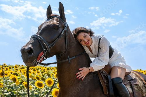 Cheerful brunette horseback riding. She is wearing traditional white dress and long black boots. Rural scene. Sunny day. Sunflower field in background.