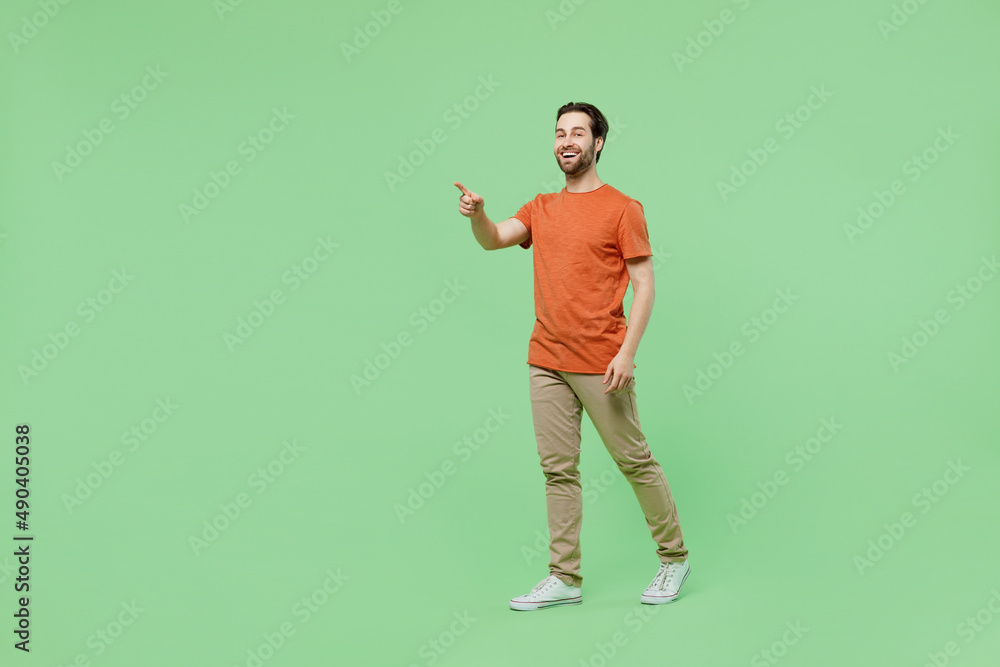 Full body side view smiling happy young man 20s wear casual orange t-shirt walking go point index finger aside on workspace area isolated on plain pastel light green color background studio portrait Full body side view smiling happy young man 20s wear casual orange t-shirt walking go point index finger aside on workspace area isolated on plain pastel light green color background studio portrait