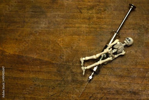 A white toy skeleton is lying on a syringe. A syringe with a skeleton on a wooden table.