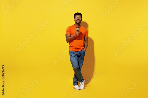 Full body young man of African American ethnicity 20s wear orange t-shirt hold takeaway delivery craft paper brown cup coffee to go isolated on plain yellow background studio People lifestyle concept.
