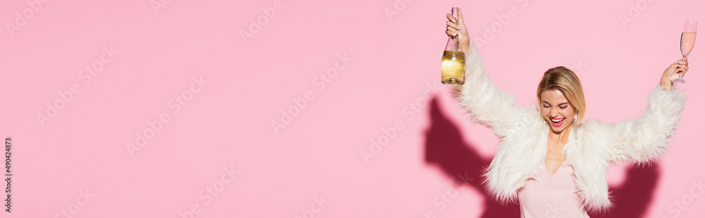 excited woman in slip dress and white faux fur jacket holding bottle of champagne and glass on pink, banner. excited woman in slip dress and white faux fur jacket holding bottle of champagne and glass on pink, banner.