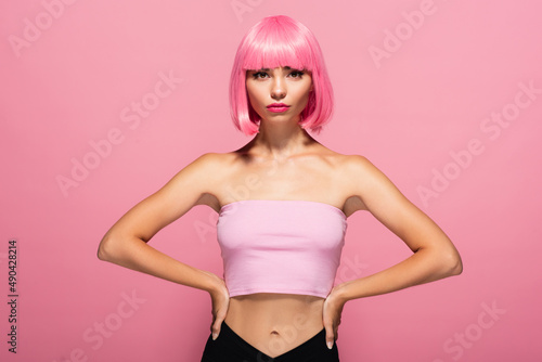 young woman with bangs and colored hair posing with hands on hips isolated on pink.