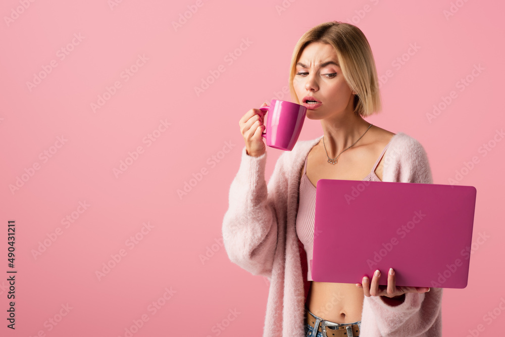 blonde woman holding mug with coffee and laptop isolated on pink.