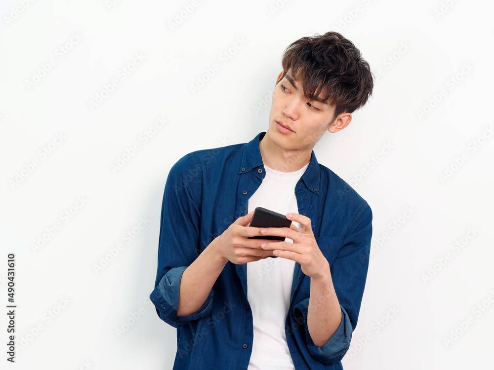 Portrait of handsome Chinese young man with curly black hair in blue shirt posing against white wall background. Looking away with mobile phone in both hands, front view studio shot. Portrait of handsome Chinese young man with curly black hair in blue shirt posing against white wall background. Looking away with mobile phone in both hands, front view studio shot.