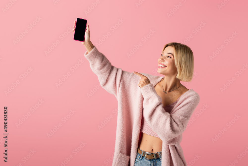 happy young woman in soft cardigan pointing at smartphone with blank screen isolated on pink. happy young woman in soft cardigan pointing at smartphone with blank screen isolated on pink.