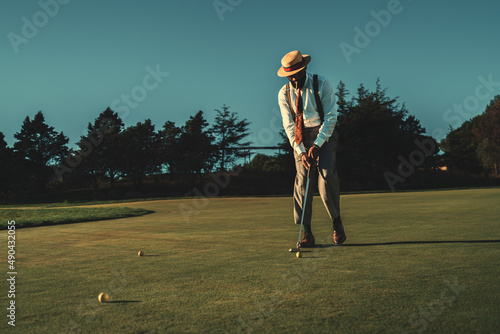 View of a fancy mature African guy in an elegant retro outfit with a straw hat and a tie hitting the ball with a club while standing on the lawn of the private golf club on a warm sunny evening